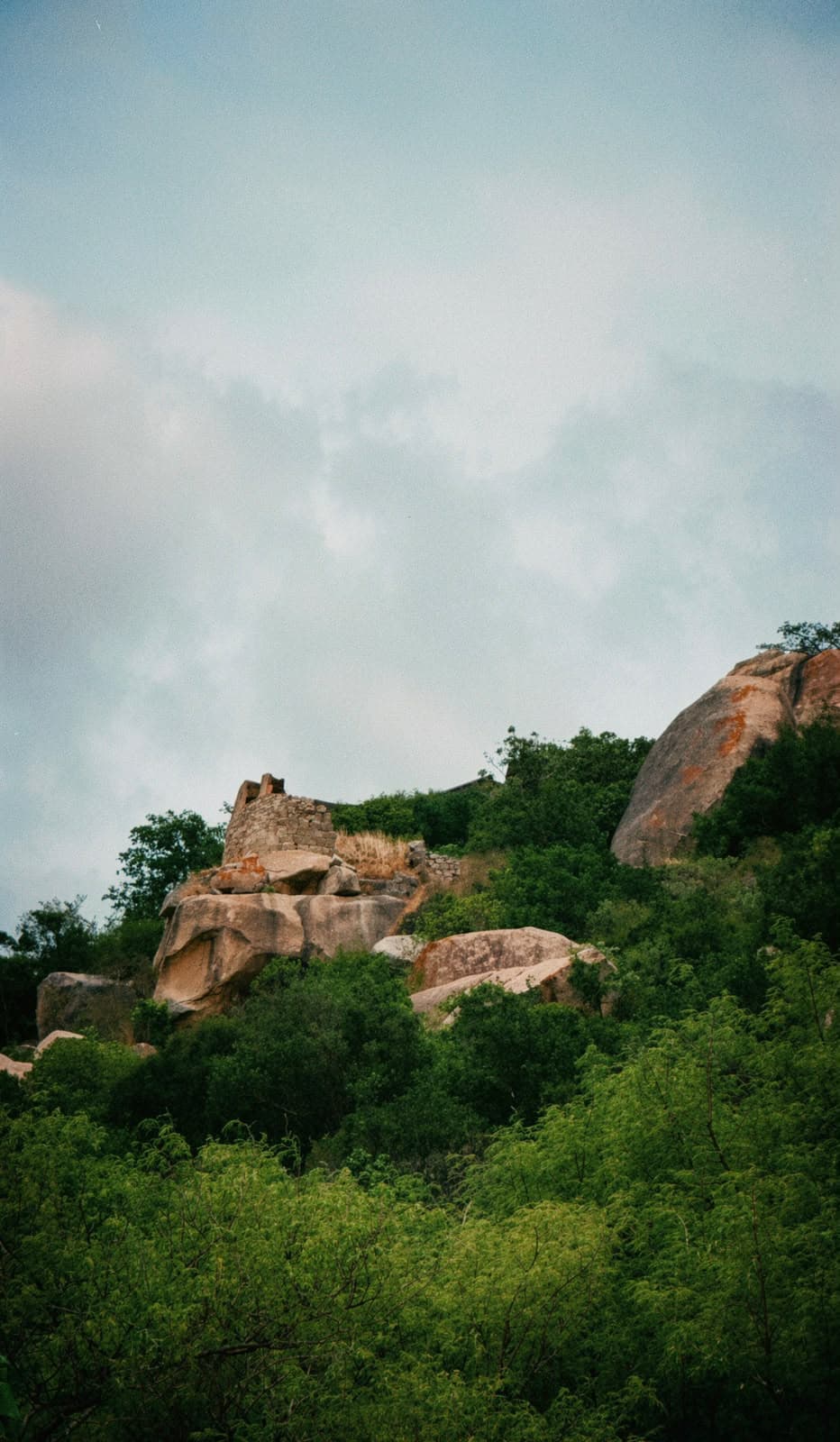 Rocky Gurramkonda hill rising above green trees beneath a cloudy sky.